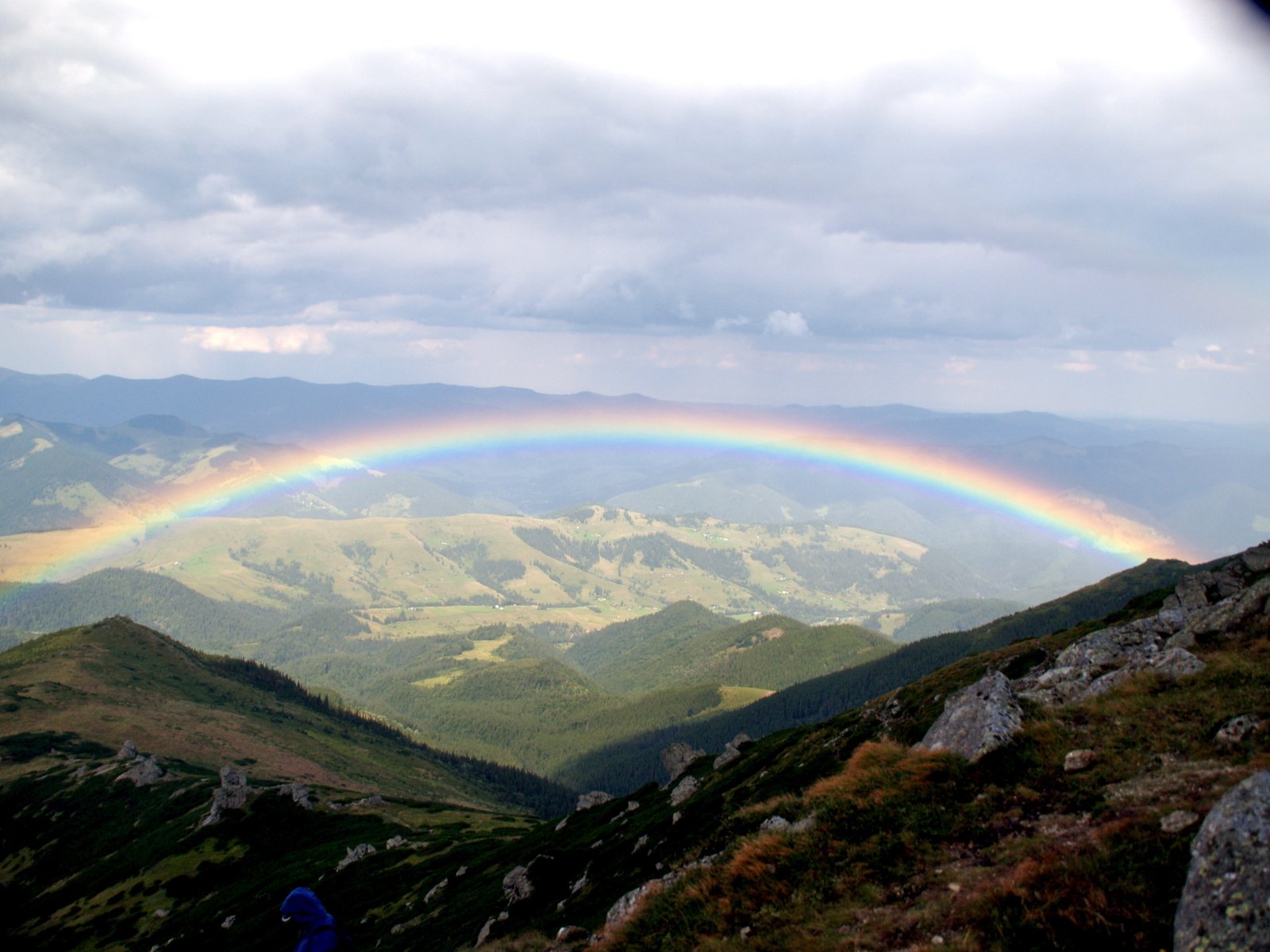 Pip Ivan in Ukraine. Photo by Dave Proffer (deepphoto on Flickr), August 2006. A rainbow arcing over two mountains with fluffy clouds above; below, green rolling hills.
